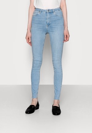 Smaltsiddende lyseblå jeans båret af en stående person, kombineret med sorte loafers og en hvid top stukket ned.