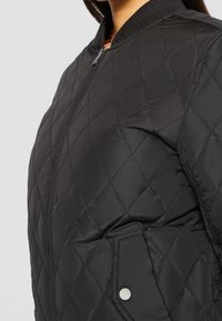 Veste bomber matelassée noire avec surpiqûres en losange, fermeture éclair, col côtelé et poche latérale à bouton-pression.