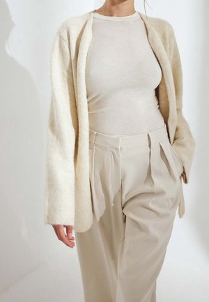 Femme portant un long cardigan beige clair sur un col roulé crème ajusté et un pantalon beige plissé taille haute.