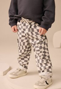 Dziecko w czarno-białych spodniaczkach w kratę, beżowych sneakersach i ciemnej, długiej koszulce stoi na beżowej okrągłej platformie.