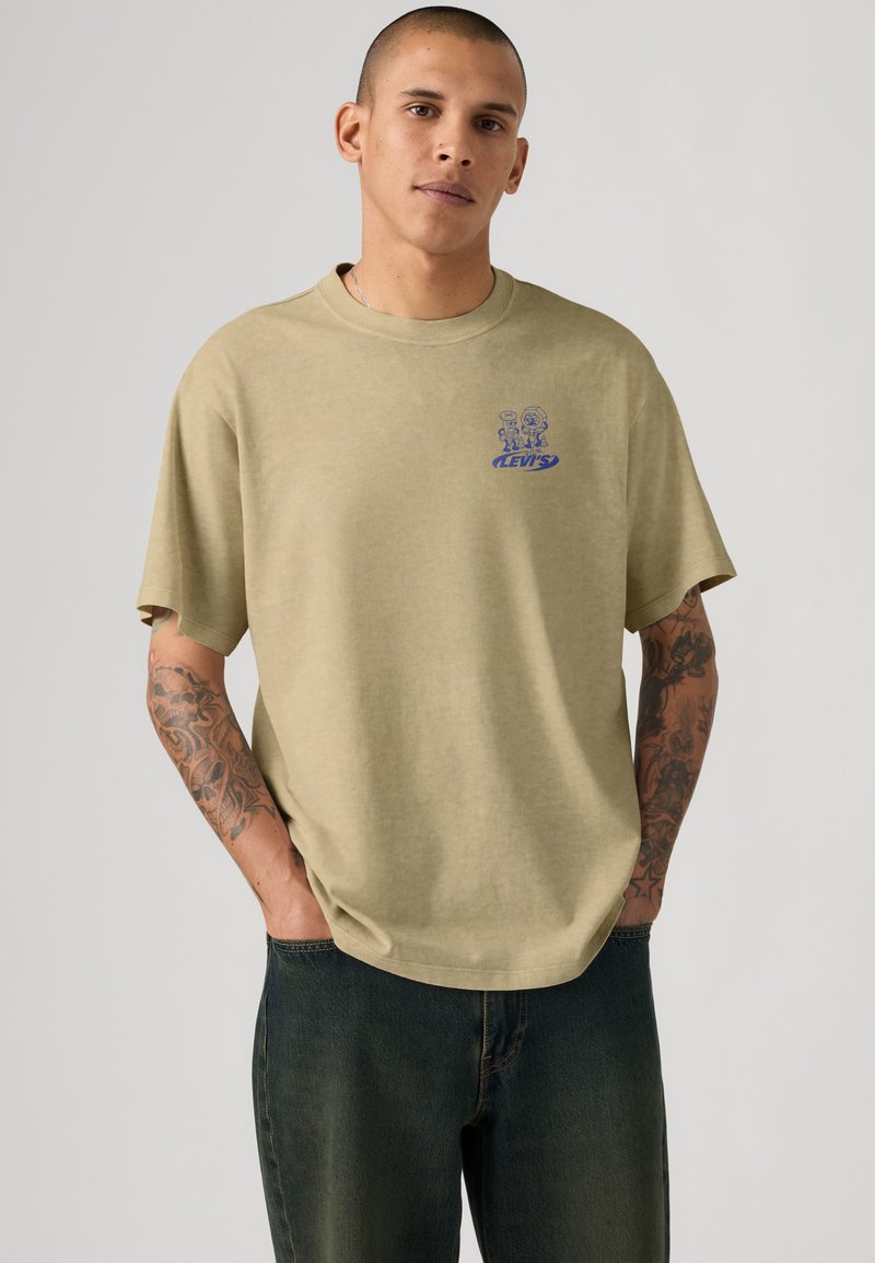 Mann mit Tattoos trägt beige Levi's T-Shirt mit kleinem blauem Aufdruck, steht mit Händen in den Taschen dunkler Jeans vor einfarbigem Hintergrund.