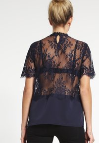 Top blu navy con retro in pizzo trasparente a fiori, maniche corte e un piccolo dettaglio a chiave sul collo. Parte anteriore in tessuto liscio.