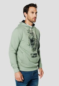 Zielona bluza z kapturem ze sznurkami, z grafiką czaszki i napisem „NIGDY NIE JEST ZA PÓŹNO”. Kieszeń typu kangur oraz ściągacze na rękawach.