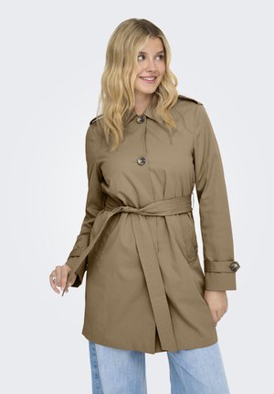 Mujer que lleva un abrigo tipo trench beige con cinturón y vaqueros azul claro, sonriendo y mirando de lado sobre un fondo liso.