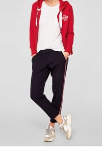 Rød hættetrøje med lynlås over en hvid T-shirt, parret med mørke bukser med røde sidestrefter og beige sneakers med detaljer.