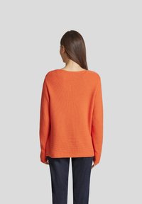 Oranger Strickpullover mit lockerer Passform, weitem Ausschnitt und gerippten Textur, kombiniert mit dunkelblauen Hosen. Rückansicht gezeigt.