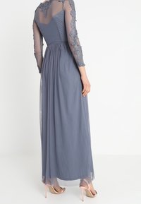 Maxiklänning i skiffergrått med långa genomskinliga ärmar och broderad spetsdetalj. Utsådd kjol med flödande tyg. Bär ljusrosa klackar.