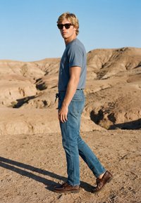Homme blond portant des lunettes de soleil, un t-shirt bleu, un jean et des chaussures marron se tient sur un terrain désertique rocailleux avec des collines arides en arrière-plan.