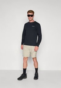 Fekete hosszú ujjú póló logóval, bézs rövidnadrág, fekete túracipő és napszemüveg. Egy férfi áll egyszerű háttér előtt.