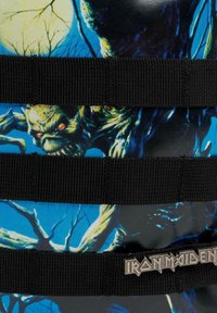 En livlig toteväska med ett monstermotiv, svarta remmar och en metallisk "Iron Maiden"-logotyp. Designen innehåller blåa och gröna nyanser med texturerade detaljer.