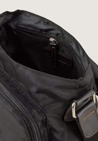 Schwarze Textiltasche mit Innenfutter, das eine mit Reißverschluss versehenen Tasche, strukturierten Stoff und Lederakzente mit genähten Details zeigt. Metallbeschläge sind sichtbar.