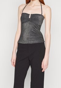 Kobieta w bezrękawnikowej, błyszczącej, ciemnoszarej bluzce na halter oraz czarnych, wysokich spodniach, przedstawiona od szyi do bioder.