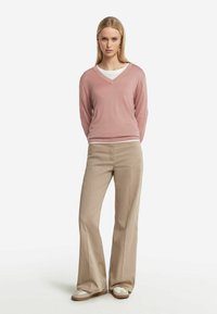 Jasnoróżowy sweter w serek wykonany z miękkiego materiału, założony na biały T-shirt. Beżowe rozszerzane spodnie i białe sneakersy dopełniają outfit.