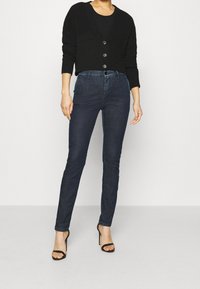 Cárdigan negro corto con tres botones, combinado con jeans ajustados azul oscuro. El modelo lleva zapatos de tacón negros con tiras. Textura de tela suave.