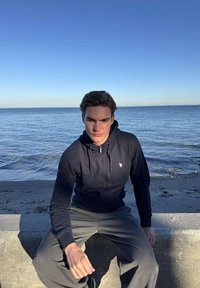 Fiatal férfi sötét kapucnis pulóverben és szürke nadrágban, aki békés tenger mellett, egy betonpadon ül, tiszta kék ég alatt.