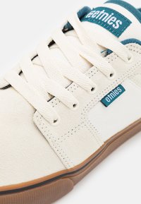 Balti sportiniai bateliai pagaminti iš šviesios zomšos ir kanvos medžiagų, su mėlynu Etnies logotipu, grietinėlės spalvos raiščių ir guminiu padu.