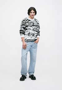 Joven de pie con un suéter de patrón abstracto en blanco y negro, jeans sueltos de color azul claro y zapatos chunky negros sobre un fondo blanco.