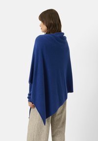 Blå, blødt strikket poncho med en draperet design, der har en høj halsudskæring og asymmetrisk kant. Materialet ser varmt og hyggeligt ud.