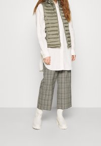 Olivově zelená puffer vesta přes bílou košili s knoflíky, doplněná šedými kostkovanými kalhotami s širokými nohavicemi a bílými kotníkovými botami s robustní podrážkou.