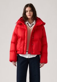 Rød puffjakke med hætte, quiltet tekstur, frontlommer, lynlåslukning og en boxy pasform, lagd over en beige sweater og en hvid skjorte.