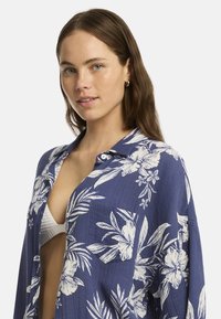 Una joven con cabello largo y castaño, vistiendo una camiseta floral azul sobre una blusa blanca, mira directamente a la cámara con una expresión neutral.