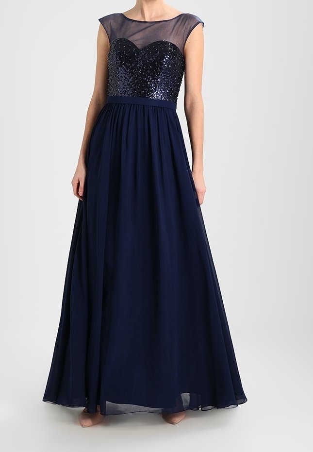 Femme portant une robe de soirée longue bleu marine avec un corsage pailleté et des manches capes transparentes, se tenant devant un fond clair et uni.