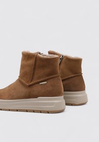Botas de tobillo de ante marrón con forro de piel de oveja, que cuentan con una cremallera trasera y una suela de goma color marrón claro con surcos texturizados. Acabado con el logo de GORE-TEX.
