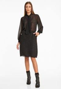 Schwarze transparente Bluse mit einem Band am Hals, getragen mit einem strukturierten schwarzen Tweedrock mit goldenen Knopfdetails und Fransensaum. Schwarze Stiefeletten.