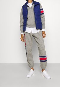 Grå sweatshirt och sweatpants-set med marinblå och röda ränder; vadderad blå väst; vit skjorta under; vita sneakers.