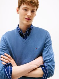 Svetlo modra v-neck pulover, položen na mornarsko modri in beli prugasti srajci. Opazna oznaka na prsi puloverja in teksturirana tkanina.