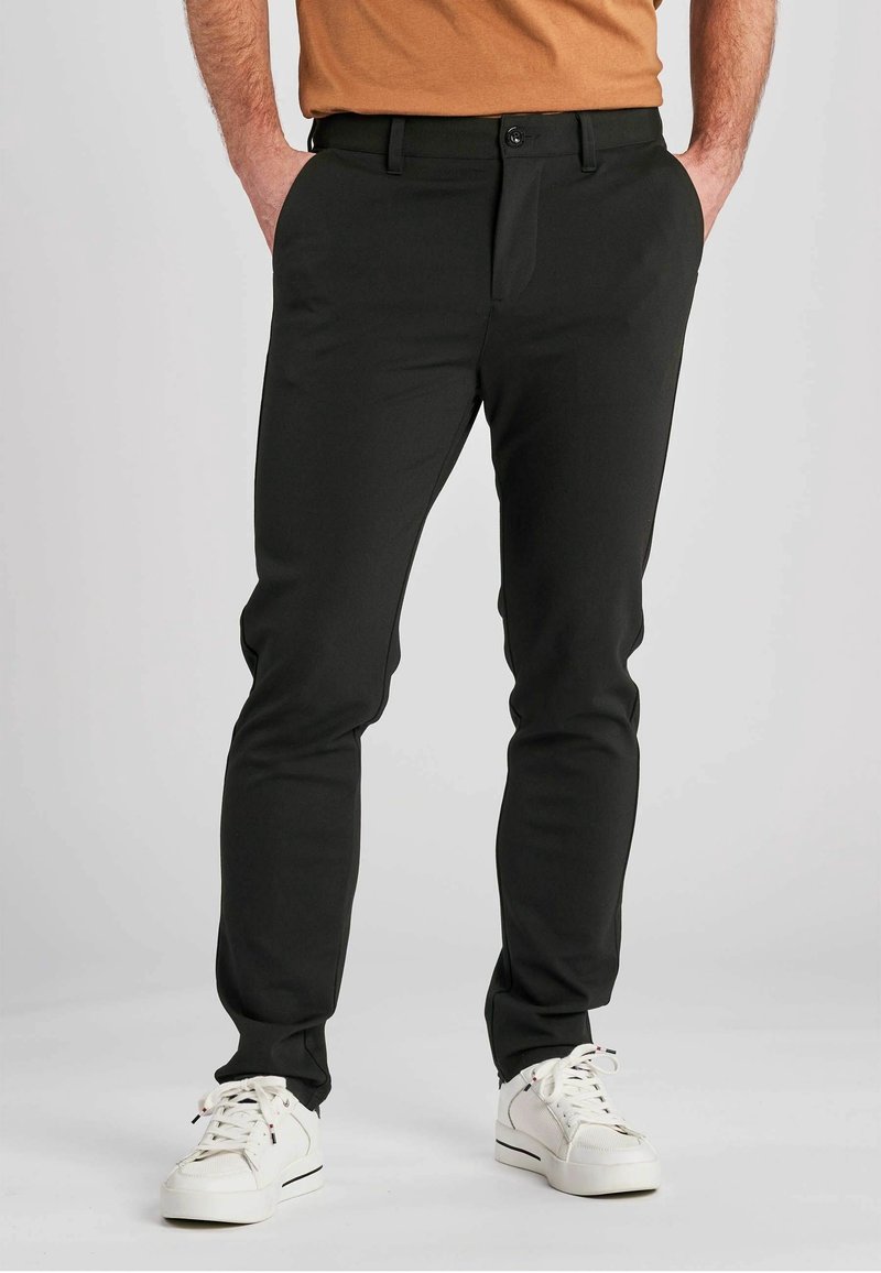 Pantalon noir ajusté en tissu lisse ; doté d'une fermeture à boutons, de passants pour ceinture et de poches latérales ; associé à des baskets blanches.