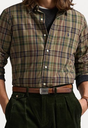 Homme portant une chemise à carreaux verte, beige et marron boutonnée sur un pull foncé, associée à un pantalon en velours côtelé vert foncé et une ceinture en cuir marron.