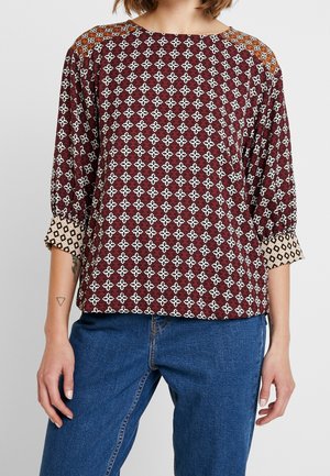 Kvinde iført mørkerød mønstret bluse med opsmøgede diamond-mønstrede manchetter og blå jeans, stående mod en ensfarvet baggrund.