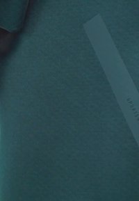 Teal fabric sports shirt with short sleeves and a smooth texture. Features a white elongated label with "SPORTS" text and a minimalist design.