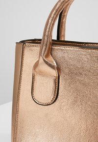 Gros plan d'un coin de sac à main métallique en or rose avec une surface texturée et une fixation de poignée cousue sur un fond clair uni.