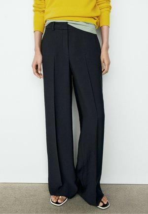 Person trägt schwarze weit geschnittene Hose, gelben Pullover, hellgrünes Hemd und schwarze Flip-Flop-Sandalen, steht vor einer weißen Wand.