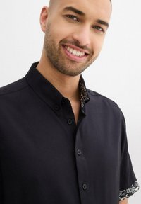 Camicia nera a maniche corte in cotone. Presenta un colletto alla francese e polsini decorativi a contrasto con un motivo decorativo.