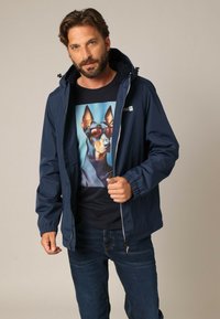 Veste coupe-vent marine avec capuche, dotée de poignets élastiques. Portée par-dessus un t-shirt noir avec un graphique coloré de chien. Jean en denim bleu.