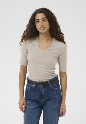 Young woman with curly hair wearing a beige V-neck short sleeve top, blue jeans, and a black belt, standing against a plain background.