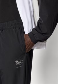 Sort sporty jakke med hvide accenter, elastiske manchetter og sidelommer; har logoet "EA7 VENTUS7" på buksebenet; letvægtsmateriale.