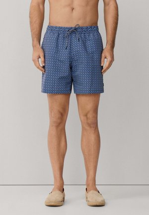 Homme portant un short de bain bleu à motifs avec un cordon de serrage, debout pieds nus sur un sol gris clair devant un mur gris uni.