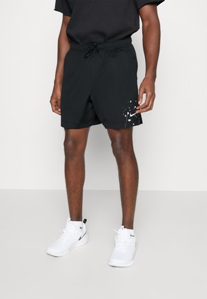 Musta urheilushortsit, joissa on joustava vyötärönauha, kiristysnauha ja alareunassa roiskemaalauskuvio, yhdistettynä valkoisiin Nike-tennareihin.