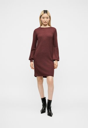 Vrouw draagt een lange mouw bordeauxrode trui jurk met opgeblazen mouwen en zwarte enkellaarsjes met spitse neuzen en metalen studs.