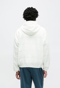 Hoodie blanc en matière douce et pelucheuse avec un motif subtil en relief. Doté d'une capuche et d'une taille élastiquée. Porté avec un pantalon bleu sarcelle.