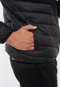 Svart puffjacka med horisontell kviltning, har en öppen sidficka. Slät textur med elastiska ärmslut och en figurnära design.
