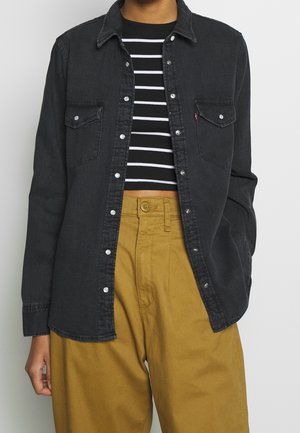 Personne portant une veste en jean noire boutonnée sur un crop top rayé noir et blanc, assortie à un pantalon taille haute jaune moutarde.