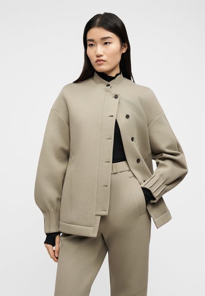 Γυναίκα με μπεζ φαρδιά σακάκι με κουμπιά και παντελόνι, με μαύρο μακρυμάνικο τοπ από μέσα, στέκεται μπροστά σε απλό φωτεινό φόντο.