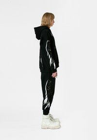 Conjunto de sudadera con capucha negra y pantalones jogger que presenta llamativos detalles en blanco y negro con forma de alas. El modelo lleva botas blancas con cordones. Textura suave y ajuste casual.