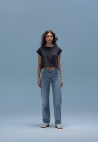 T-shirt preta cropped, jeans de cintura alta azul claro e ténis brancos. Modelo está em pé contra um fundo azul claro.