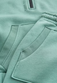 Weicher grüner Stoff in Nahaufnahme zeigt elastischen Bund und Seitentaschen an lässigen Jogginghosen und passendem Oberteil.